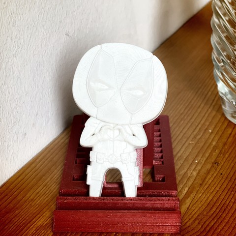 A 3D printed figure resembling a stylized Deadpool character stands atop a red platform, beside a clear glass bottle, against a white wall.