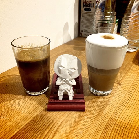 A half-empty glass of dark liquid next to a latte in a clear glass, with a stylized Deadpool figure seated in front of them on a wooden surface. Boxes of food and water bottles are in the background.