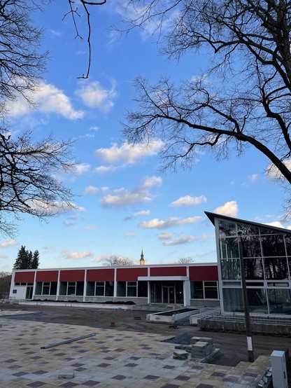 Modern building with red accents and large glass windows, under a blue sky with sparse clouds, flanked by bare trees.