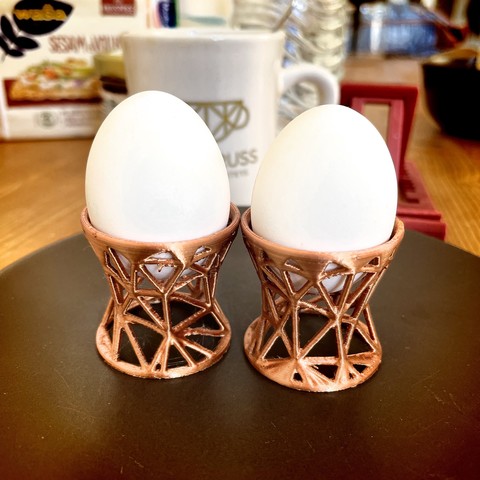 Two white eggs in copper geometric egg cups on a brown plate with kitchen items in the background. The the egg cups are 3D printed