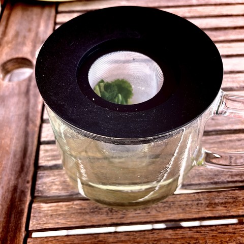 A glass teapot with a black lid, containing fresh peppermint tea, on a wooden table.