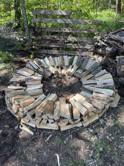 A circular stack of chopped firewood with a wooden fence in the background.