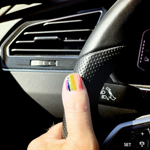 Close-up of a thumb with rainbow-colored nail polish holding a car steering wheel.
