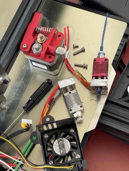 Close-up of 3D printer components, including a stepper motor with a red extruder, a fan, wires, screws, and other metal parts on a metal surface.