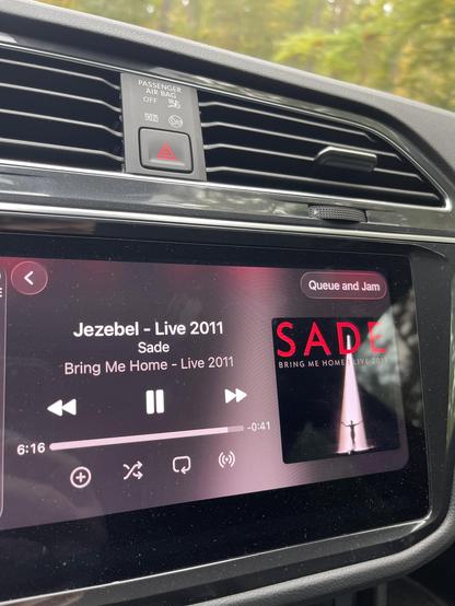 The image shows a car's dashboard display featuring a music player interface. It is currently playing "Jezebel - Live 2011" by Sade from the album "Bring Me Home - Live 2011." The display includes playback controls and
