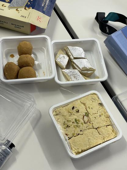 An assortment of Indian sweets is displayed on a table. There are three round, golden-brown sweets in one container, pieces of silver-coated barfi (a type of fudge) in another, and a rectangular block of white and cream-colored sweet with pistachios 