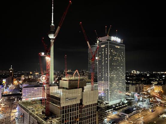 Nighttime cityscape featuring the Berlin television tower and construction cranes. Nearby buildings are illuminated, showcasing a bustling urban environment.