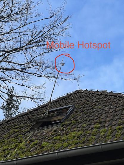 A roof with moss-covered tiles features an open window and a tall pole with a mobile hotspot device attached. The background shows bare tree branches against a blue sky, with the words "Mobile Hotspot" prominently displayed in red.