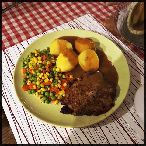 Ein grüner flacher Teller auf dem Kartoffeln, buntes Mischgemüse und ein Schnitzel (ohne Panade) angerichtet sind. Die Kartoffeln und das Schnitzel sind teilweise mit einer braunen Soße bedeckt. Der Teller steht auf einer weißen Unterlage mit wenigen unregelmäßigen roten Streifen. Die Unterlage wiederum steht auf einem rot/weiß karierten Tischtuch. 