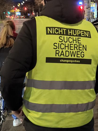 Eine Person mit schwarzer Jacke und signalgrüner Warnweste. Steht offenbar an einer Ampel als Radfahrer. Auf der Warnweste steht in schwarzer Schrift: Nicht Hupen! Suche sicheren Radweg. 