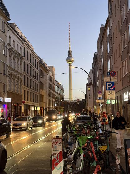 A busy city street with cars, bikes, and pedestrians. The Berlin TV Tower stands tall in the background under a clear blue sky.