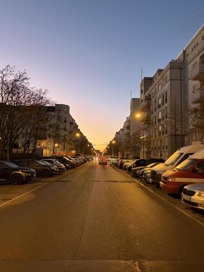 A city street lined with parked cars and buildings under a blue sky with a yellow sunrise.