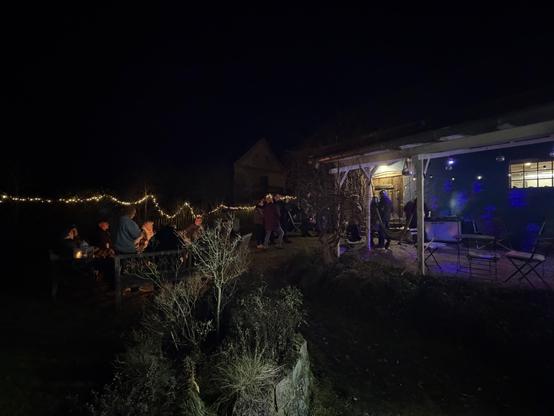 Menschen sitzen an einem Tisch mit Kerzen unter Lichtern im Freien. Im Hintergrund ist ein Gebäude mit blauen Lichtern.