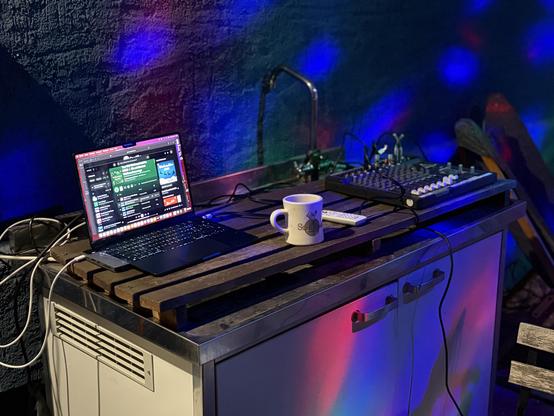 Ein kleiner Schrank auf dem ein Laptop mit Musikapp steht. Rechts daneben ein Mischpult. Der Hintergrund eine raue Wand auf der einige bunte Lichtflecken zu sehen sind.