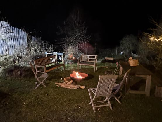 Holzstühle um ein Feuer im Garten bei Nacht, umgeben von Stühhlen und einem Tisch mit einer Korb.