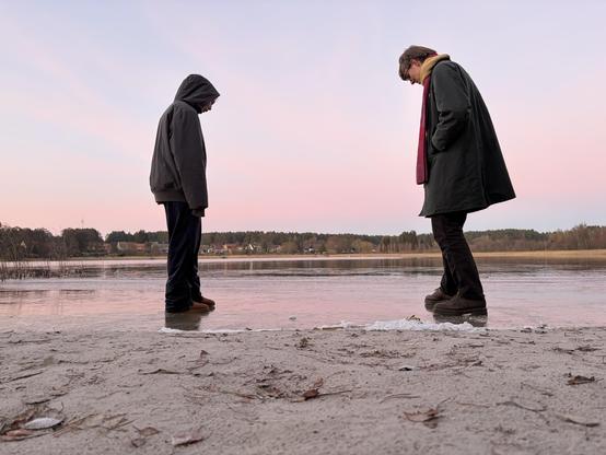 Two persons standing on top of the water of a small lake. Apparently the water at the shore is frozen to hold the weight. Both people are wearing thick winter clothes. The right person is also wearing a scarf. The sky is clear and the beginning sunset turning the edge pink from blue already. The photo is taken from a very low angle. 