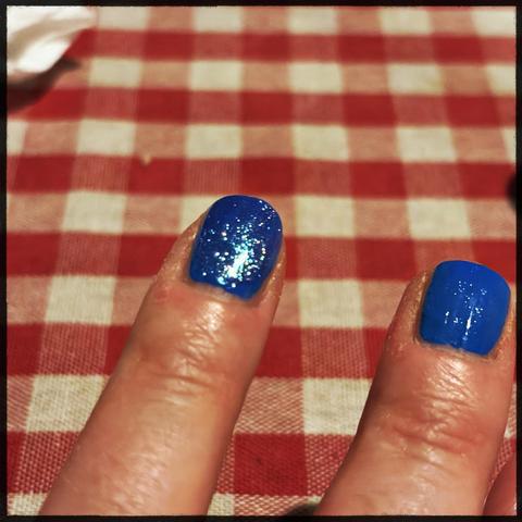 Image shows two fingers with bright blue glitter nail polish. The background features a red and white checkered tablecloth. The left finger has glitter coating on top of the color. On the right it's just light reflecting 