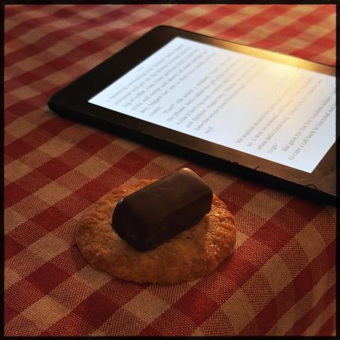 A chocolate-covered cookie and an e-reader on a red and white checkered tablecloth.