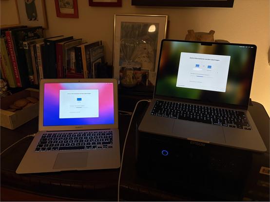 Two MacBook Air laptops on a desk, one older and one newer, both displaying setup screens. Books and a framed picture in the background.