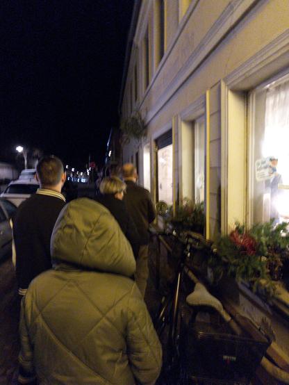 Menschen stehen in einer Schlange vor einem hell beleuchteten Fenster mit Dekorationen in der Nacht.