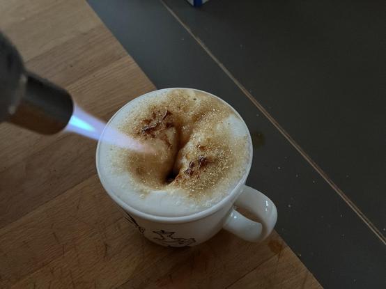 A white mug with a black design holds a frothy coffee, with steam rising from it on a wooden surface. A kitchen torch is caramelizing the sugar in the coffee with a blue flame.
