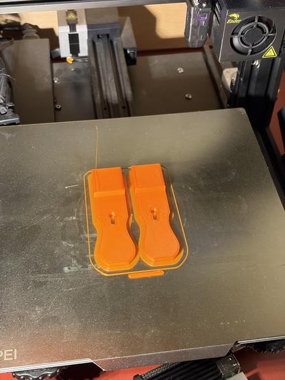 Two plastic latches on a metal sheet print bed of a 3D printer. The pieces are made of orange plastic.