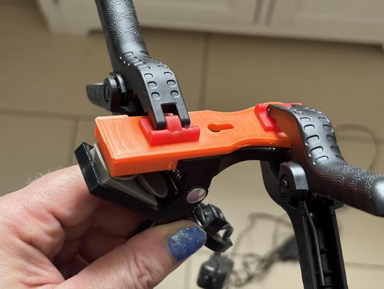 Two clamps are holding an orange plastic latch to a black  clamp of an IKEA LED lamp. The orange plastic piece seems being glued to the black clamp and hold in place by the two other plastic clamps. A left hend is holding the setup. The thumb has blue nail polish.