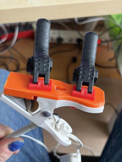 Two clamps are holding an orange plastic latch to a white  clamp of an IKEA LED lamp. The orange plastic piece seems being glued to the white clamp and hold in place by the two other plastic clamps. A left hend is holding the setup. The thumb has blue nail polish.