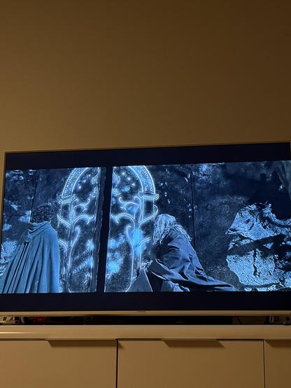 Zwei Personen stehen vor einem blauen Tor in einem Wald. Eine Person ist in einen Umhang gehüllt, die andere kniet und berührt das Tor.