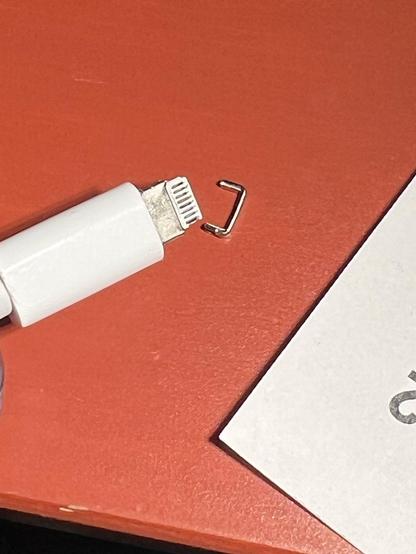 A white lightning power cable for Apple iPhones und a red surface. Right of the tip is a tiny u-shaped metal part. Looking almost like a tiny stapler needle. That's the tip of the lightning cable plug that's broken off and got stuck in the iPhone port.