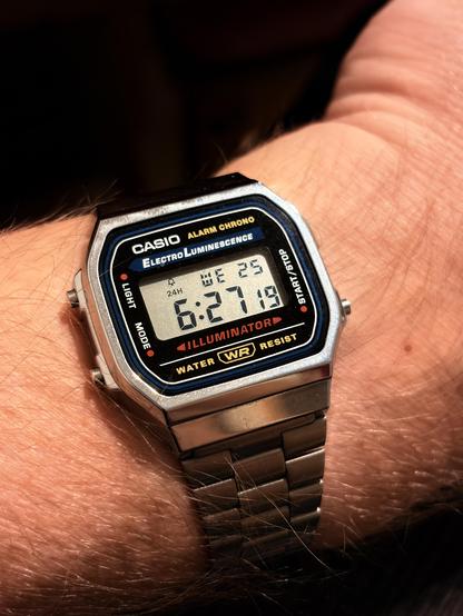 A man's wrist with a classic silver CASIO watch displaying the time 6:27:19. The watch has a digital display and a metal band.
