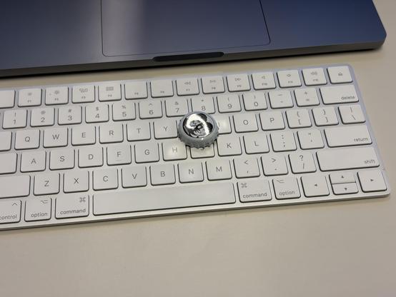 A white keyboard with a silver bottle cap on the "k" key, next to a laptop.