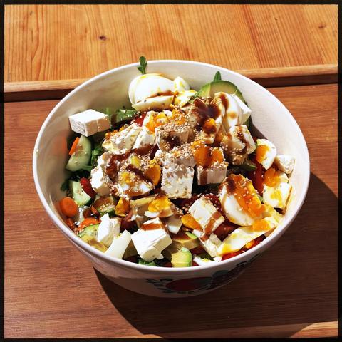 A white bowl with a salad of vegetables, tofu, and eggs, topped with sesame seeds and brown sauce, on a wooden surface.
Ein bunter Salat mit Tofu, Eiern, Gurken, Karotten und Paprika, garniert mit Sesam und einer braunen Sauce, in einer weißen Schälchen.
