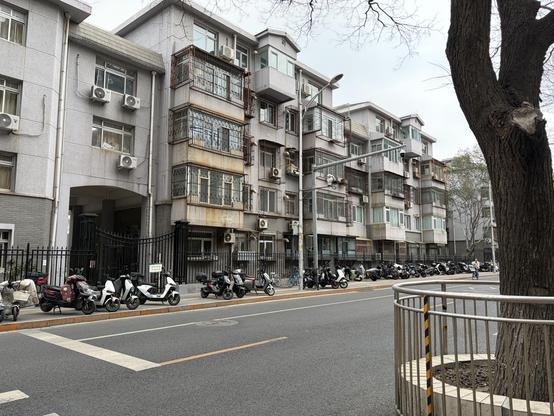 A row of multi-story buildings with balconies and air conditioners. Scooters parked on the sidewalk. A tree in the foreground.