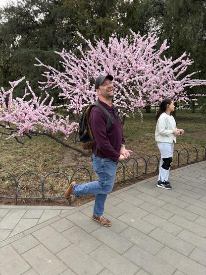 A man in a maroon sweater and jeans poses playfully in front of a pink flowering tree, while a woman stands nearby.
