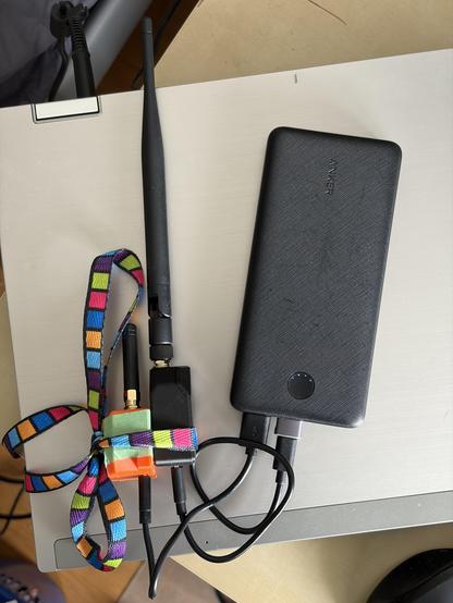 Two tiny radio devices in 3D printed casings. One in black, one in orange. The black one has a long antenna where the small one has only a short antenna. The two are tied together with a colourful shoelace. Both are connected via short black USB cables to a black Anker power bank. The whole setup rests on an closed old HP laptop .