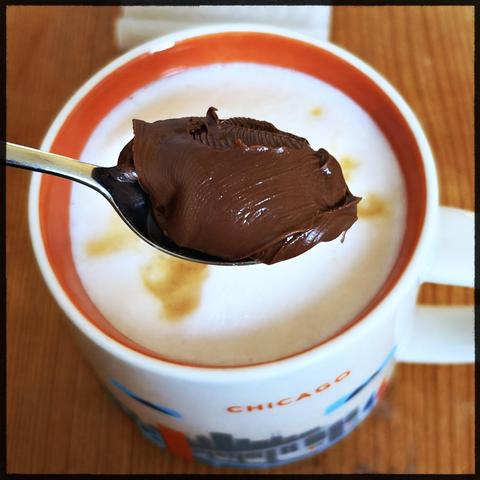 Ein Löffel Nutella über einer Tasse heißem Kaffee auf einem Holzbrett.