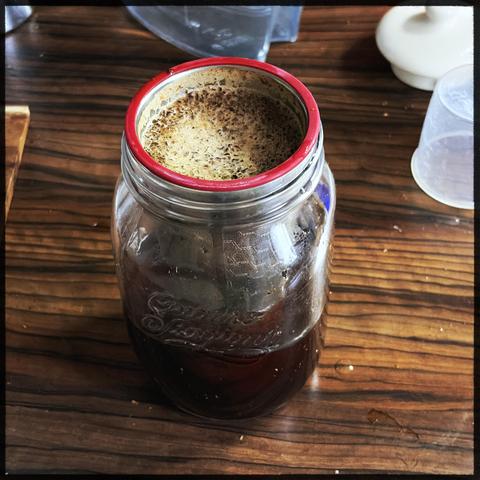 A mason jar with a metal filter tube inside. The filter tube is filled with coarse coffee grounds and water. The brim of the filter is red rubber. The jar sits on a dark wooden tabletop.