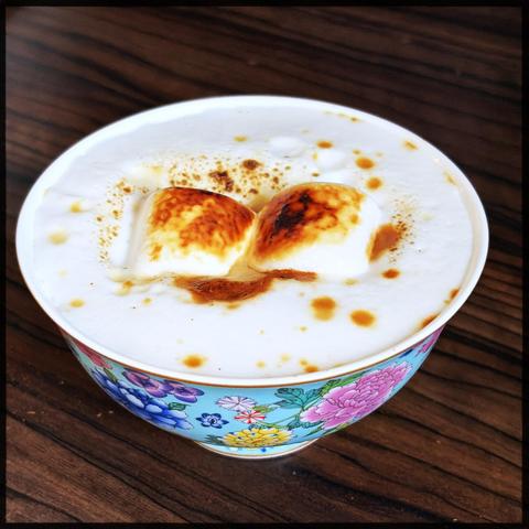 A white bowl with a floral pattern holds a creamy beverage (coffee) topped with toasted marshmallows on a wooden surface.