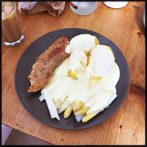 Ein Teller mit Spargel, Sauce Hollandaise und Fleisch, dazu ein Glas Milchkaffee auf einem Holztisch.