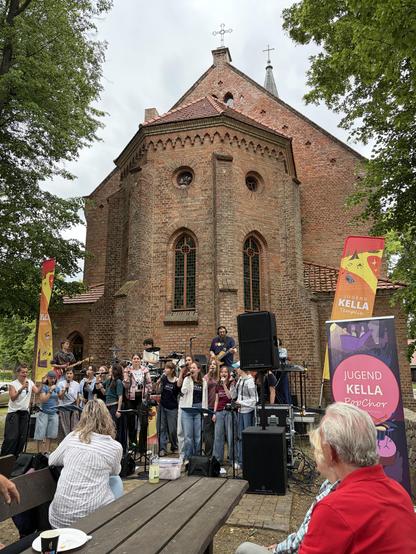 Chor vor einer Backsteinkirche, mit Musikern und Publikum, Banner "Jugend Kella PopChor", sonniger Tag.