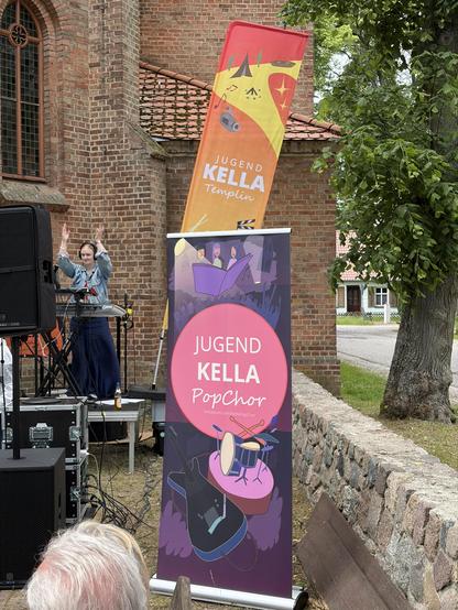 Jugend Kella PopChor tritt vor einem Backsteingebäude auf. Banner mit Musikinstrumenten, Frau am Keyboard, Publikum im Freien.