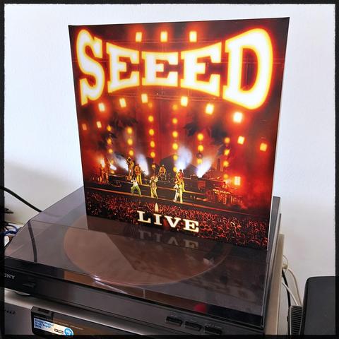 Vinyl-Platte von "Seed" auf einem Sony-Plattenspieler, Cover zeigt Band auf Bühne mit roten Lichtern und "Live" Schriftzug.