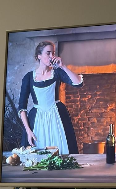 Eine Frau in einem historischen Kleid trinkt aus einem Glas Rotwein, während sie in einer rustikalen Küche Gemüse schneidet.