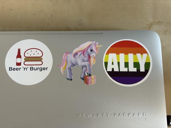 A laptop with three stickers: a unicorn, a rainbow "ALLY," and a "Beer 'n' Burger" logo.