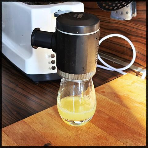 A small glass with lemon juice at the bottom. On top a black portable espresso maker from Wacaco. The picoPresso is a black cylinder with a piston sticking out one side. The setup rests on a wooden board on a countertop