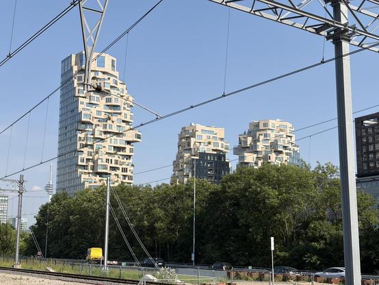 Three unique, stacked buildings with balconies and trees in front, under a clear blue sky with power lines.