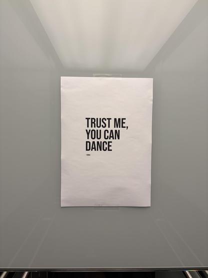 A white poster with black text reading "Trust me, you can dance. --- Vodka" is taped to a white wall in an elevator