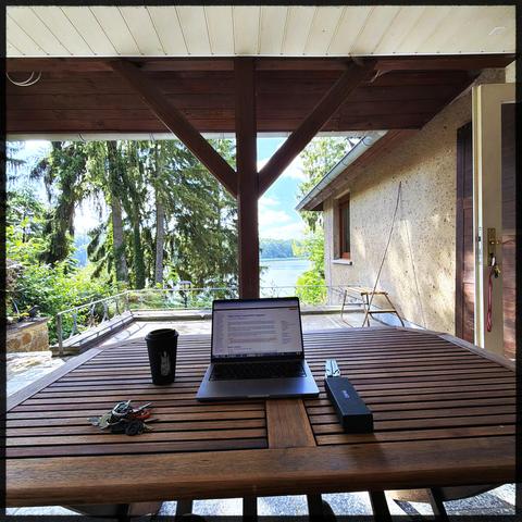 A wooden table on a terrace near a house. In the background trees and a lake. On the table is a laptop , a black coffee mug, a leatherman multitool and a glasses box.