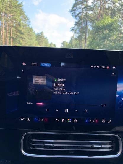 Ein on-Board Entertainment System eines Autos zeigt den Spotify Player. Es wird aktuell der Song "Lunch" von Billie Eilish gespielt. Durch die Windschutzscheibe sieht man Wald und ein Stück Himmel mit ein paar Wolken.